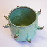 Three-quarter overhead view of empty handmade dark clay spiked vessel; bright seafoam-teal glaze with subtle blue sheen at the rim; curved brown-tipped protruding spikes; dark unglazed clay base.