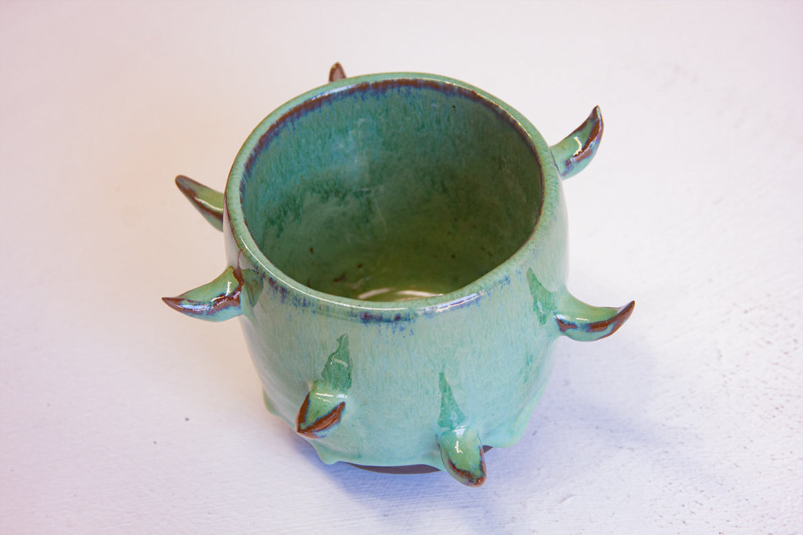 Three-quarter overhead view of empty handmade dark clay spiked vessel; bright seafoam-teal glaze with subtle blue sheen at the rim; curved brown-tipped protruding spikes; dark unglazed clay base.