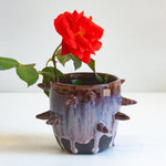 Side view of handmade dark clay spiked vessel holding a red rose; deep plum-burgundy glaze with pale lavender drips running to the dark unglazed base; protruding pointed clay spikes around the cylindrical body.