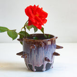 Side view of handmade dark clay spiked vessel holding a red rose; deep plum-burgundy glaze with pale lavender drips running to the dark unglazed base; protruding pointed clay spikes around the cylindrical body.