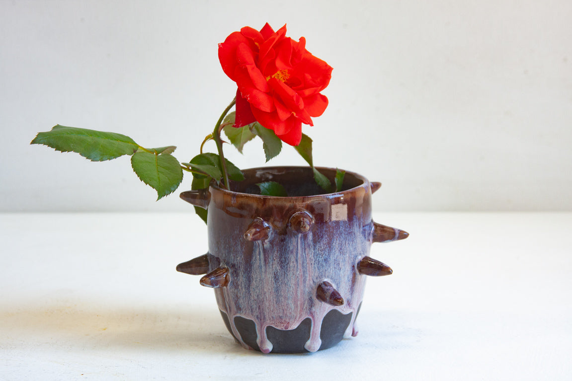 Side view of handmade dark clay spiked vessel holding a red rose; deep plum-burgundy glaze with pale lavender drips running to the dark unglazed base; protruding pointed clay spikes around the cylindrical body.