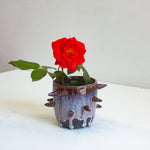 Side view of handmade dark clay spiked vessel holding a red rose; deep plum-burgundy glaze with pale lavender drips running to the dark unglazed base; protruding pointed clay spikes around the cylindrical body.