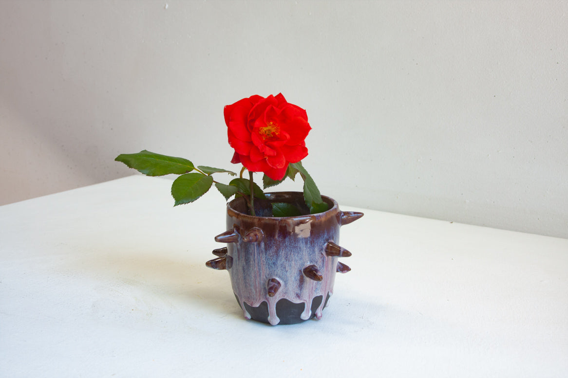 Side view of handmade dark clay spiked vessel holding a red rose; deep plum-burgundy glaze with pale lavender drips running to the dark unglazed base; protruding pointed clay spikes around the cylindrical body.