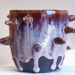 Front-facing side view of handmade dark clay spiked vessel against a pink background; deep plum-burgundy glaze with pale lavender-white drips running to the unglazed dark base; multiple protruding clay spikes around the body.