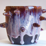 Front-facing side view of handmade dark clay spiked vessel against a pink background; deep plum-burgundy glaze with pale lavender-white drips running to the unglazed dark base; multiple protruding clay spikes around the body.