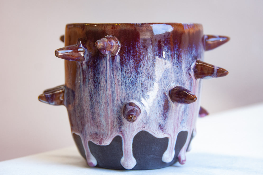 Front-facing side view of handmade dark clay spiked vessel against a pink background; deep plum-burgundy glaze with pale lavender-white drips running to the unglazed dark base; multiple protruding clay spikes around the body.