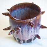 Three-quarter overhead view of empty handmade dark clay spiked vessel; cylindrical form with deep plum-burgundy glaze fading to pale lavender with white drips; multiple protruding pointed spikes; dark unglazed base.