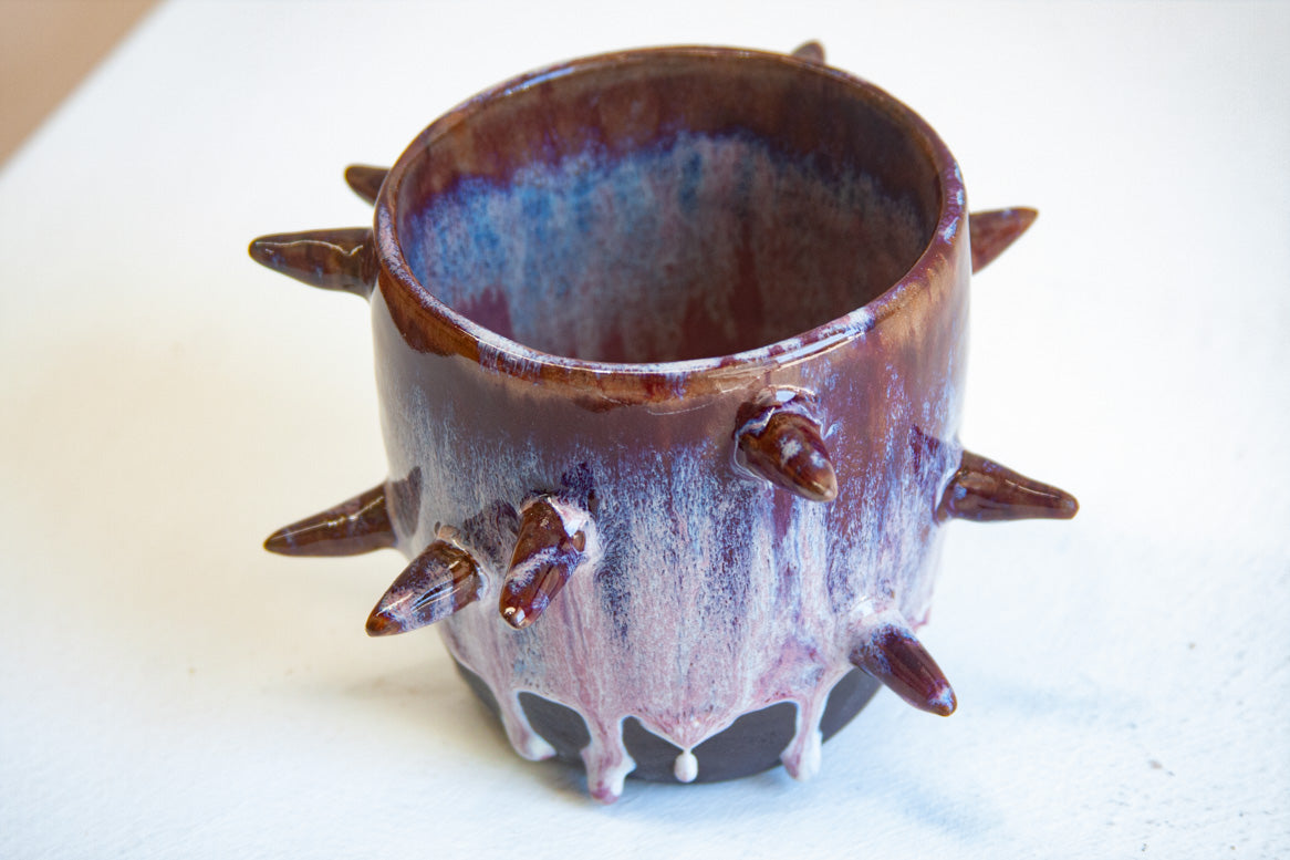 Three-quarter overhead view of empty handmade dark clay spiked vessel; cylindrical form with deep plum-burgundy glaze fading to pale lavender with white drips; multiple protruding pointed spikes; dark unglazed base.
