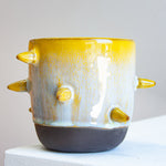 Front-facing side view of handmade dark clay spiked vessel on a white plinth; bright golden-yellow glaze fading to speckled pale blue-grey; protruding pointed yellow spikes; prominent unglazed dark clay base.