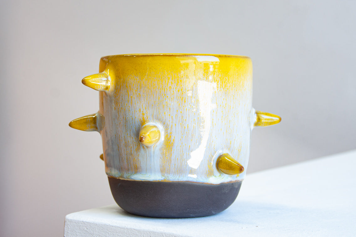 Front-facing side view of handmade dark clay spiked vessel on a white plinth; bright golden-yellow glaze fading to speckled pale blue-grey; protruding pointed yellow spikes; prominent unglazed dark clay base.