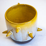 Three-quarter overhead view of empty handmade dark clay spiked vessel; bright golden-yellow glaze dripping to pale speckled blue-grey; six protruding pointed yellow spikes; dark unglazed clay base.