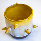 Three-quarter overhead view of empty handmade dark clay spiked vessel; bright golden-yellow glaze dripping to pale speckled blue-grey; six protruding pointed yellow spikes; dark unglazed clay base.