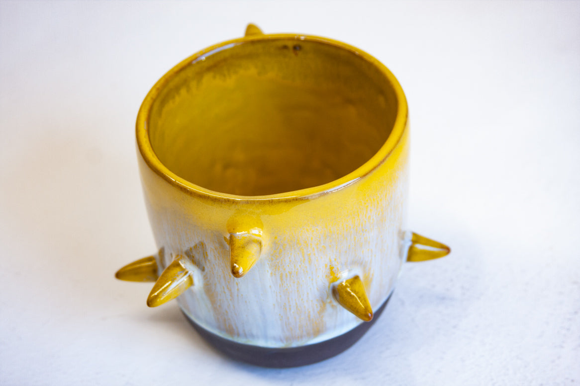 Three-quarter overhead view of empty handmade dark clay spiked vessel; bright golden-yellow glaze dripping to pale speckled blue-grey; six protruding pointed yellow spikes; dark unglazed clay base.