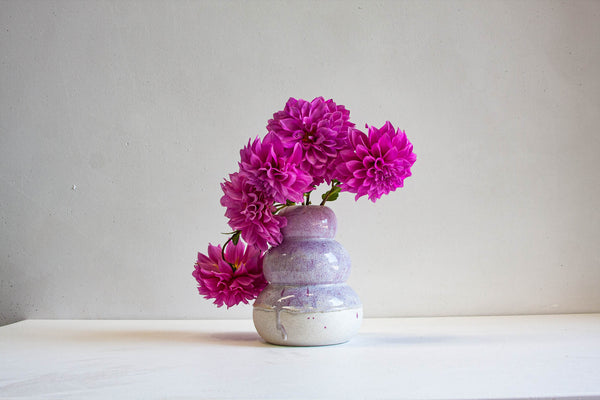 Lifestyle shot of handmade ceramic large smoosh vase with two stacked rounded lobes in soft lilac-lavender glaze over white speckle, unglazed white speckled stoneware base, filled with magenta dahlias