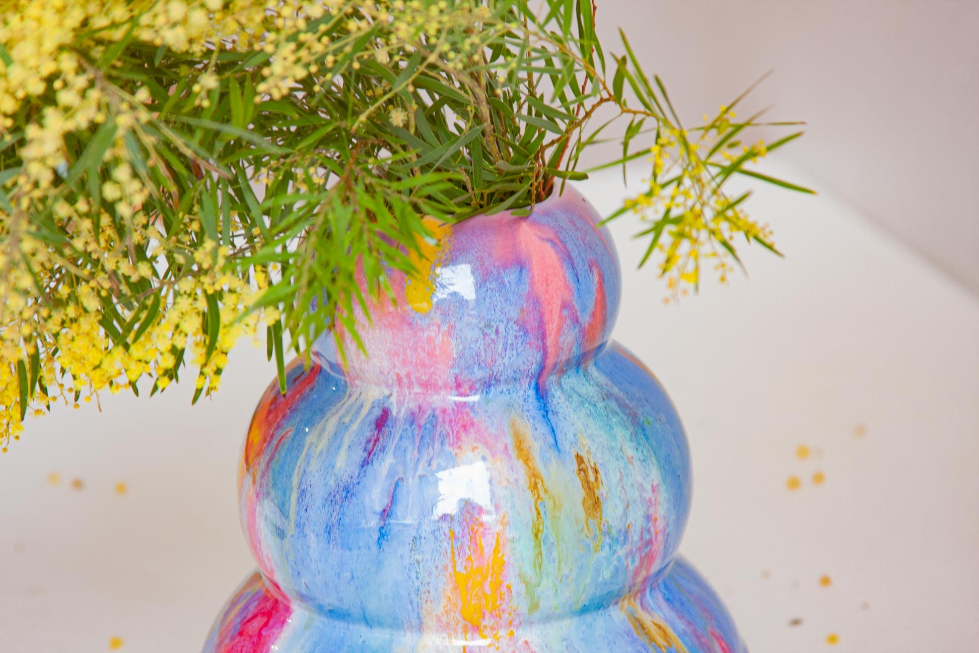 A close-up of a glossy ceramic vase with a marbled swirl pattern of blue, pink, purple, and orange, filled with green foliage and yellow flowers against a light background.