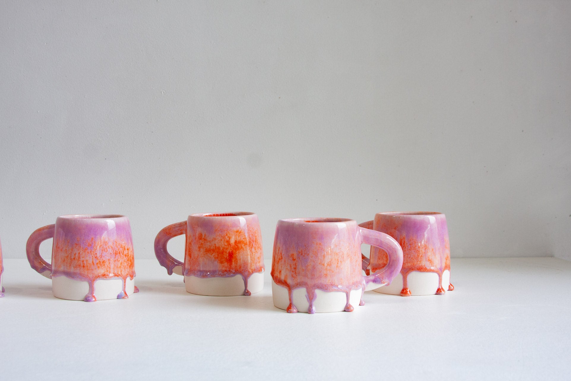 Four small ceramic mugs with a pink and orange drip-glaze finish, arranged in a row against a plain white background.
