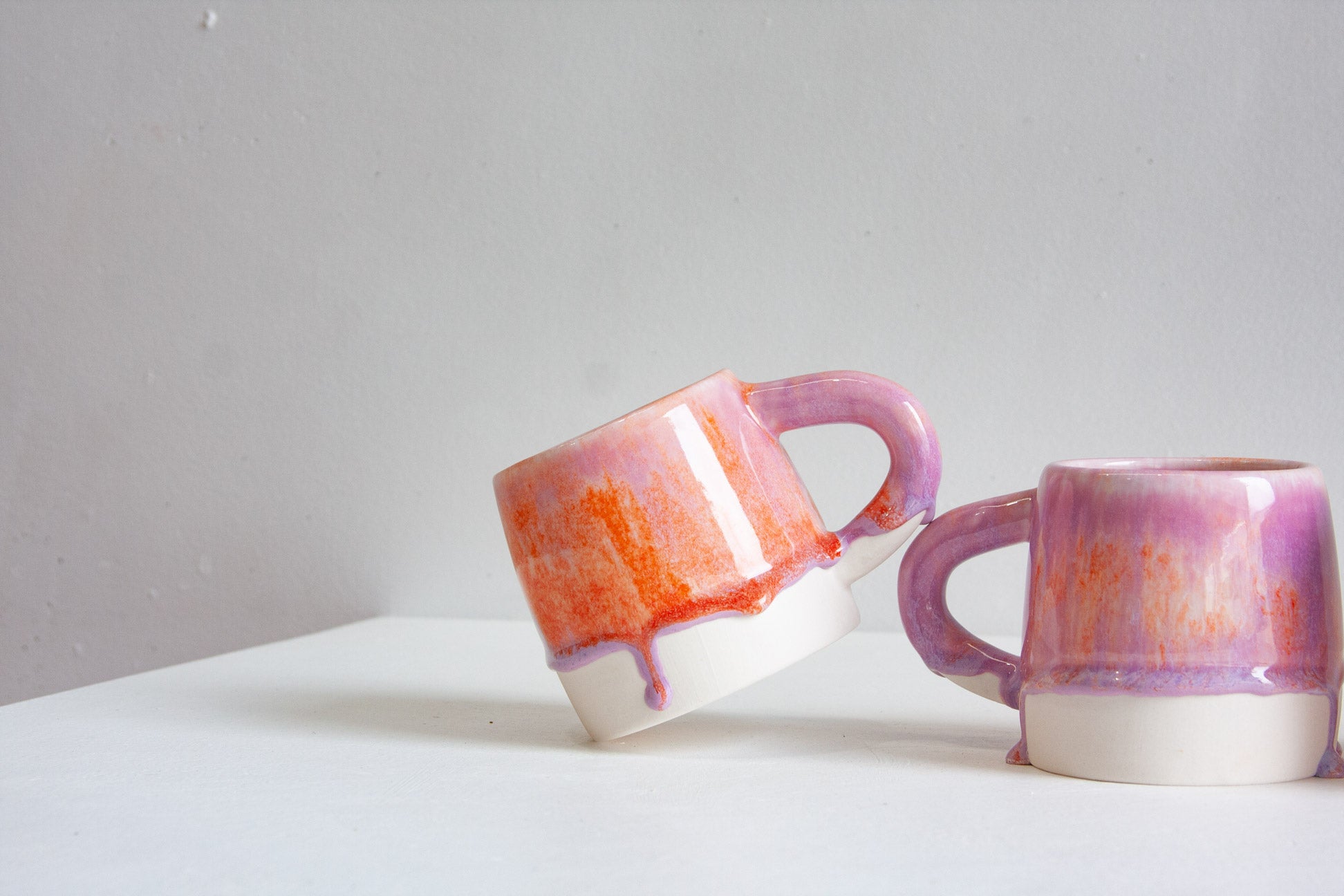 Two handmade ceramic mugs with a glossy finish, featuring a white base and a purple and orange dripping glaze, displayed on a white surface against a light gray background.