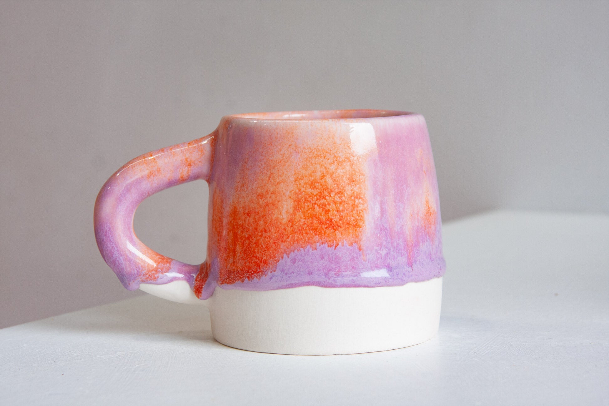 A handmade ceramic mug with a dripping glaze design, featuring a white base, a purple and orange gradient upper body, and a purple glazed handle, set against a neutral gray background.