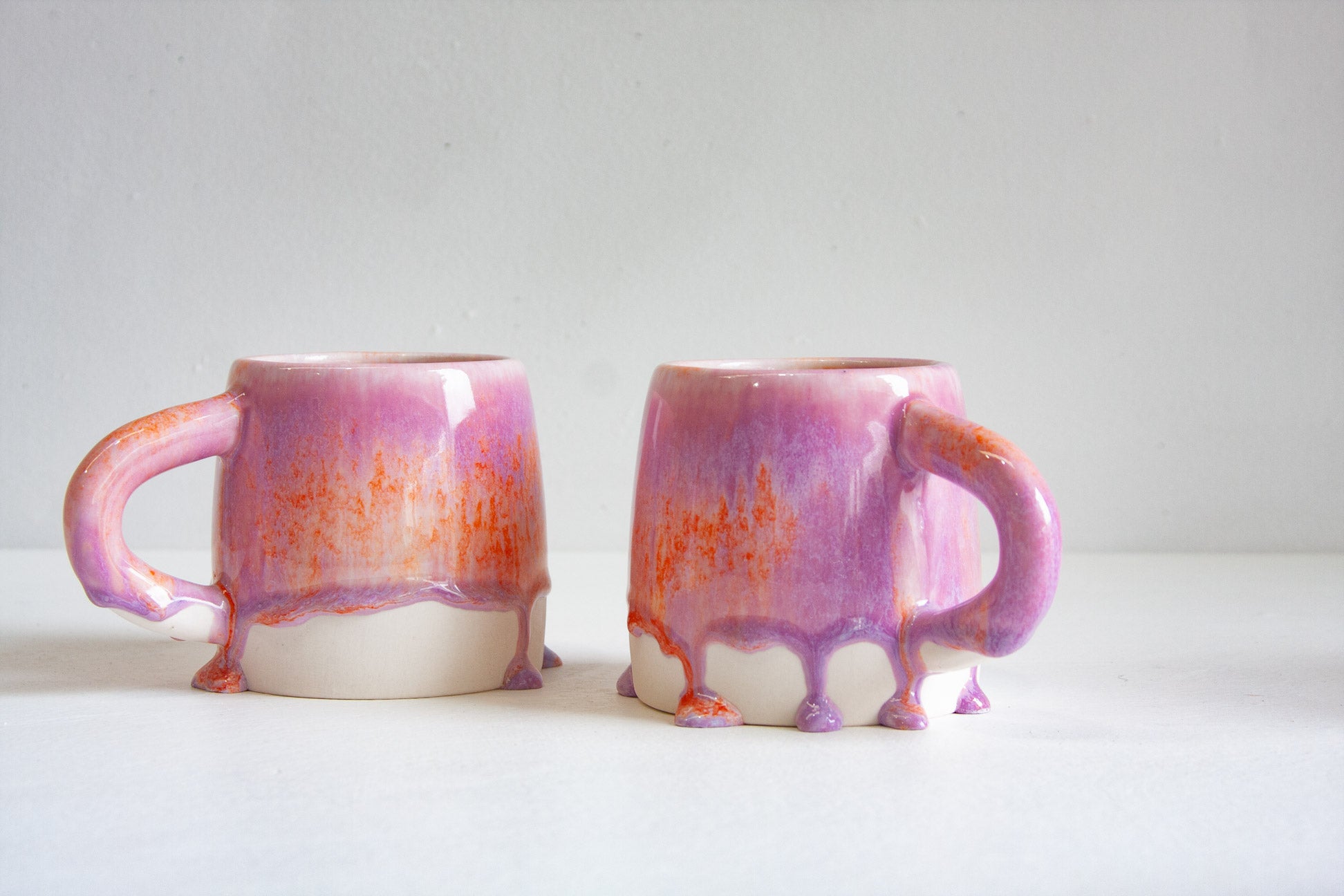Two ceramic mugs with a dripping glaze design in shades of purple, orange, and white, set against a plain light gray background.