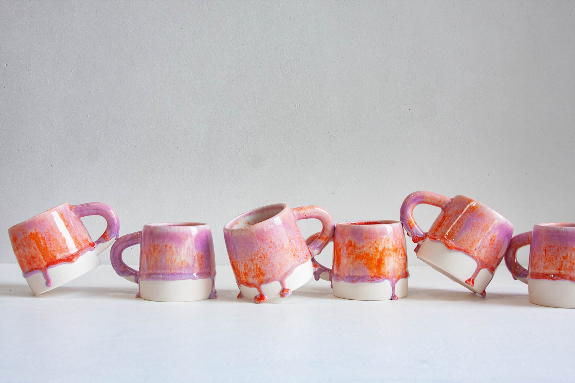 Five small ceramic mugs with dripping orange and purple glaze, arranged in a row against a light gray background.