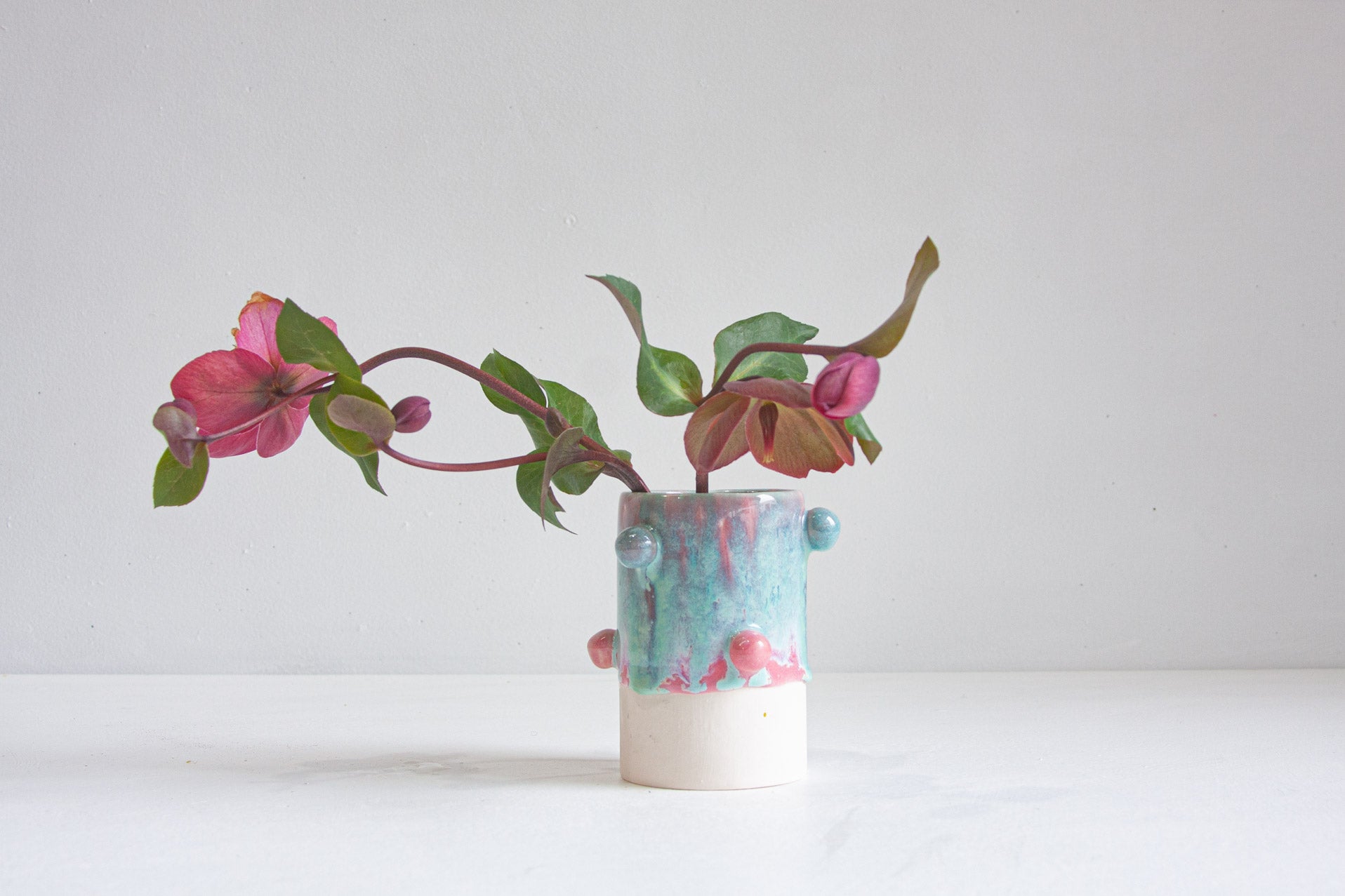 A small handmade ceramic vase with a marbled turquoise, pink, and white glaze, holding a flowering green branch with pink blossoms, set against a plain light gray background.