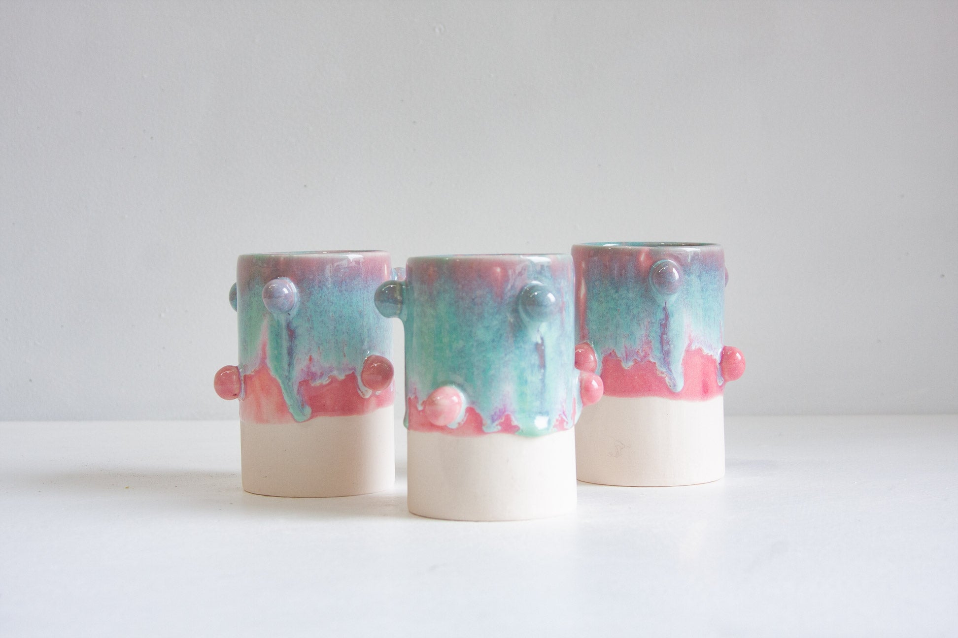 Three handmade ceramic planters with a dripping pastel glaze in pink, teal, and cream, featuring small raised decorative knobs, displayed on a white surface against a light gray background.