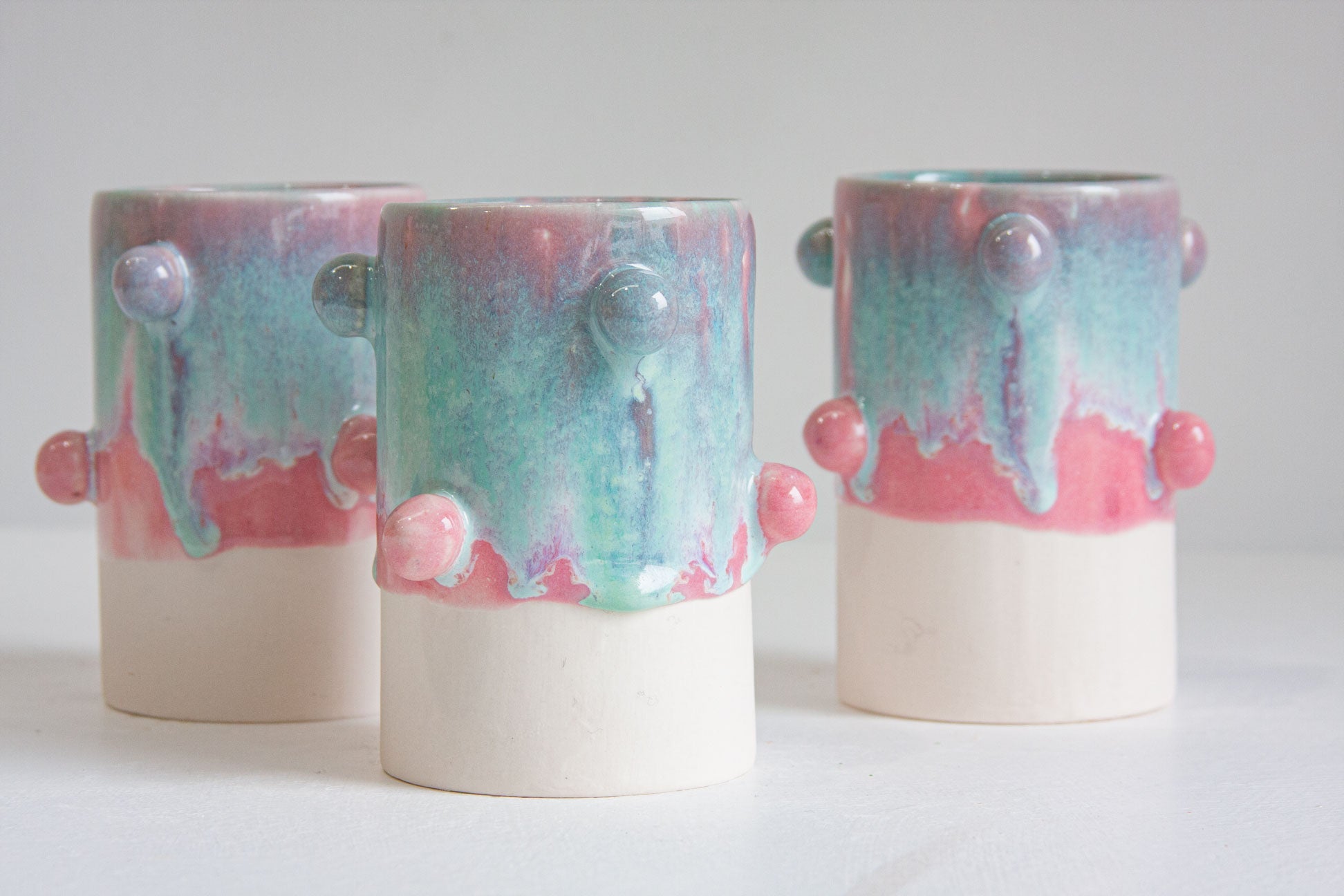 Three handmade ceramic vases with a dripping pastel glaze in pink, teal, and purple, featuring textured raised details and a solid cream base, displayed on a light gray surface.