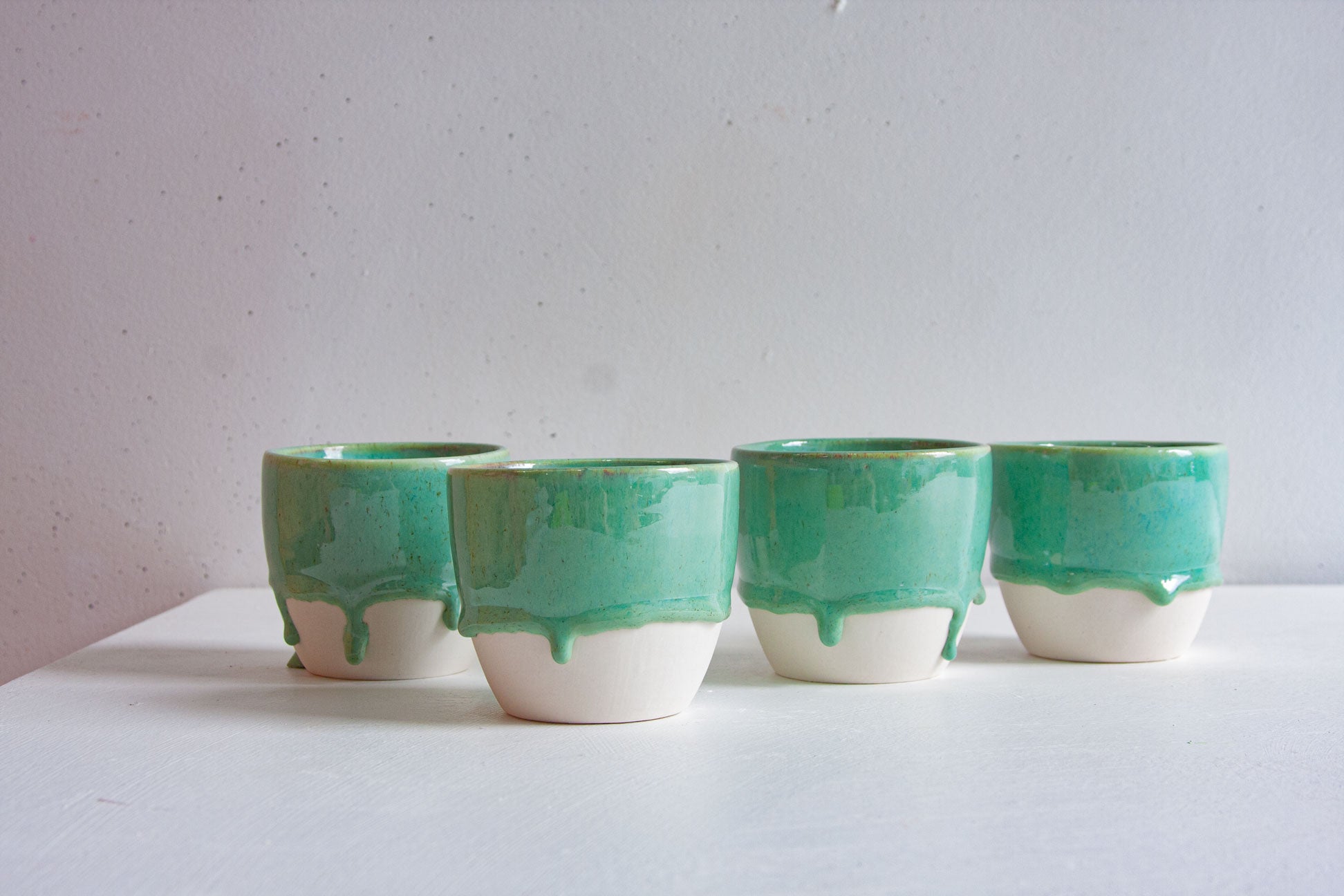 Four handmade ceramic cups in a row, dip-glazed in soft blue-green that runs into drips above the bare white clay base.