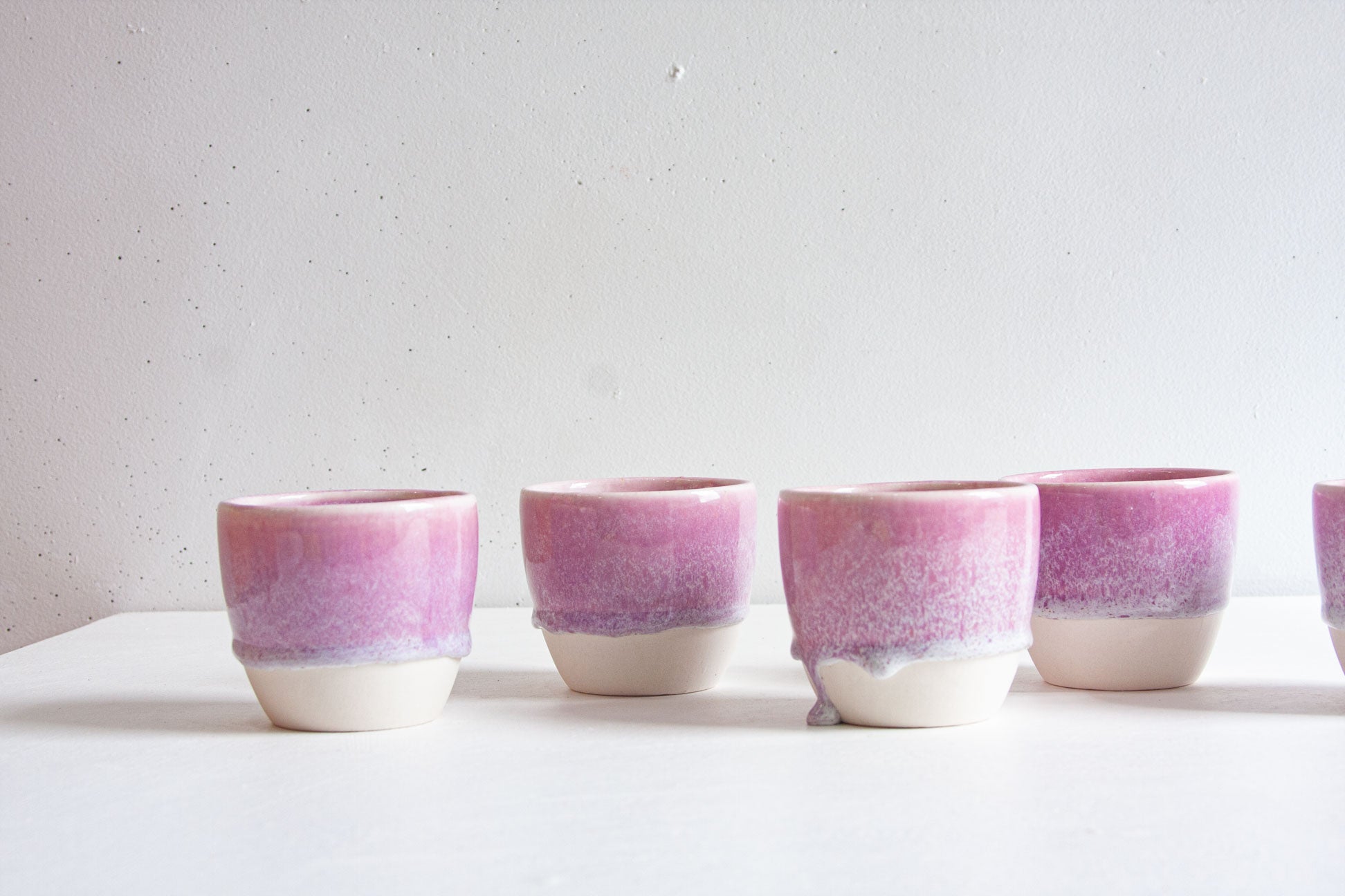 Five handmade ceramic cups in a row, dip-glazed in light purple breaking into white speckles above the bare white clay base, on a plain light background.