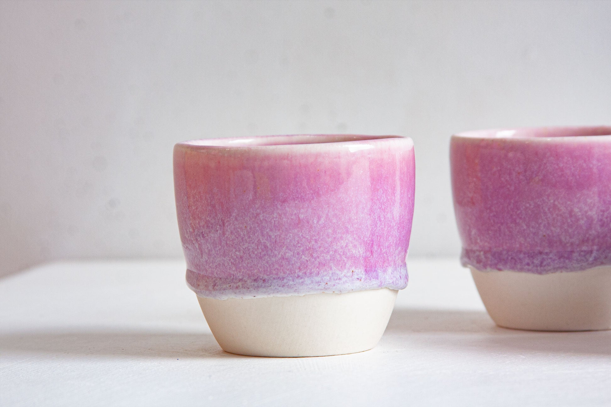 Close-up of two handmade ceramic cups, the light purple and white glaze ending in a soft wavy line above the bare white clay base.