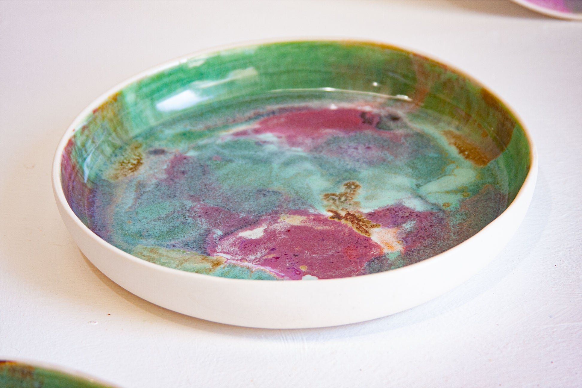 A shallow round ceramic plate with a white rim and a marbled interior featuring swirls of green, teal, magenta, and gold glaze, photographed on a white surface.