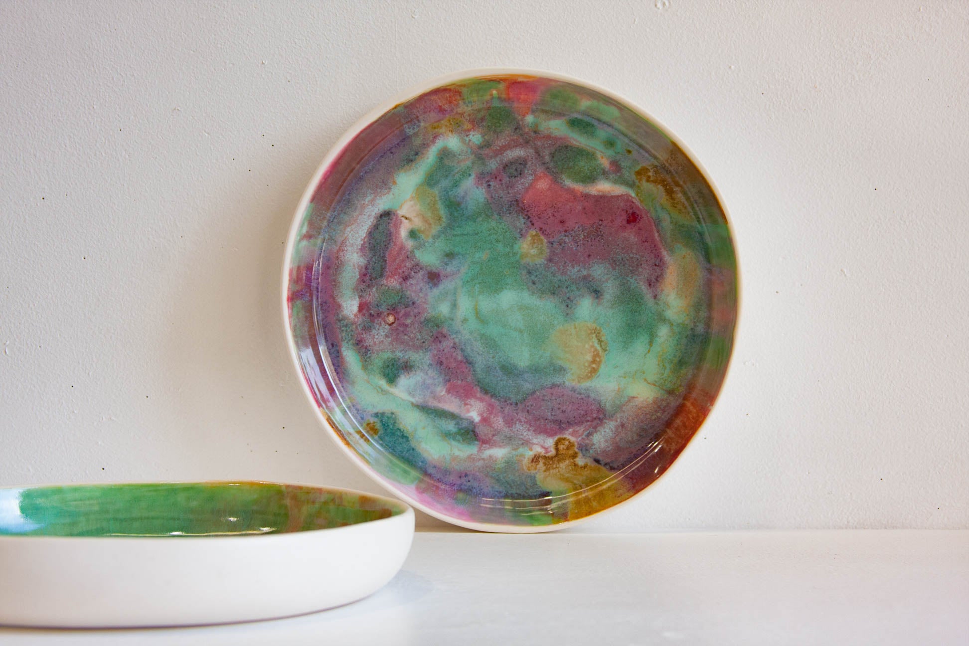 A round ceramic plate with a marbled, hand-painted glaze in shades of green, pink, purple, and gold, displayed against a plain off-white wall with another matching plate partially visible in the foreground.