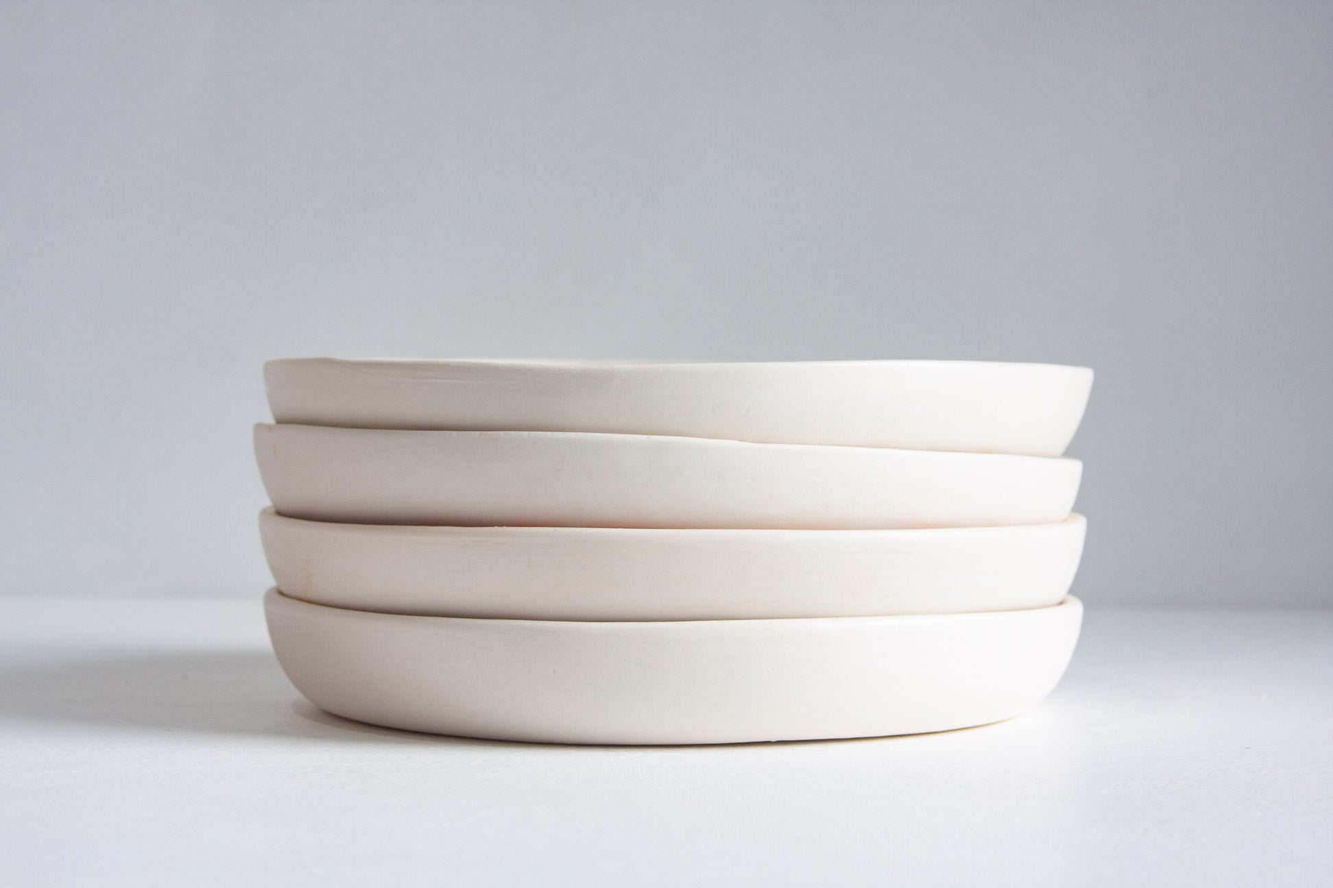 A stack of four shallow, wide ceramic plates in a warm off-white or cream color, photographed against a neutral light gray studio background.