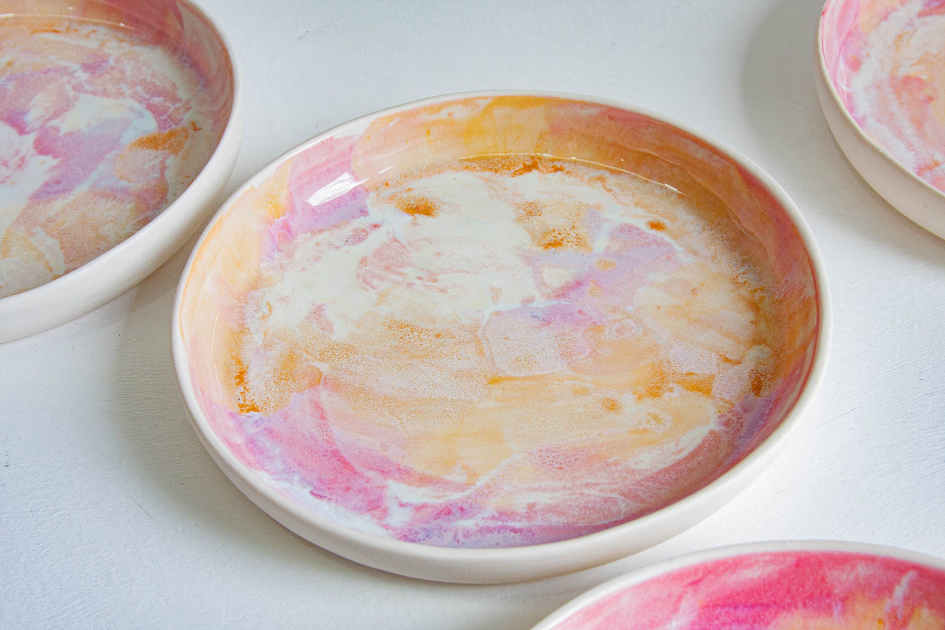 A close-up of a shallow round ceramic plate with a marbled swirl pattern in pink, orange, and cream, with other similar plates partially visible around it on a white surface.