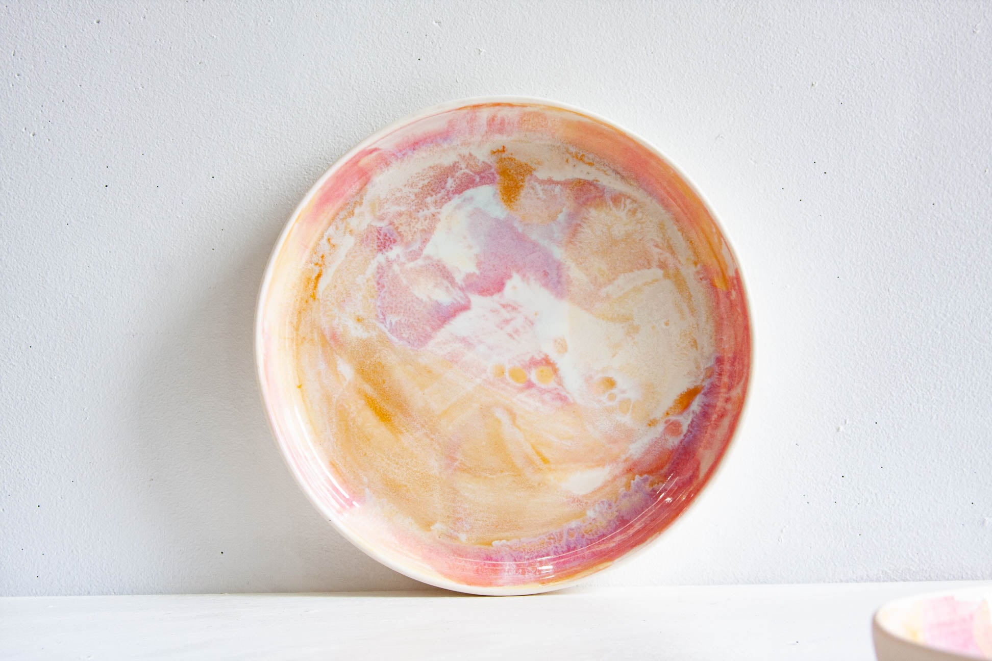 A round ceramic plate with a marbled, hand-painted design in pink, peach, orange, and cream, set against a plain white wall.