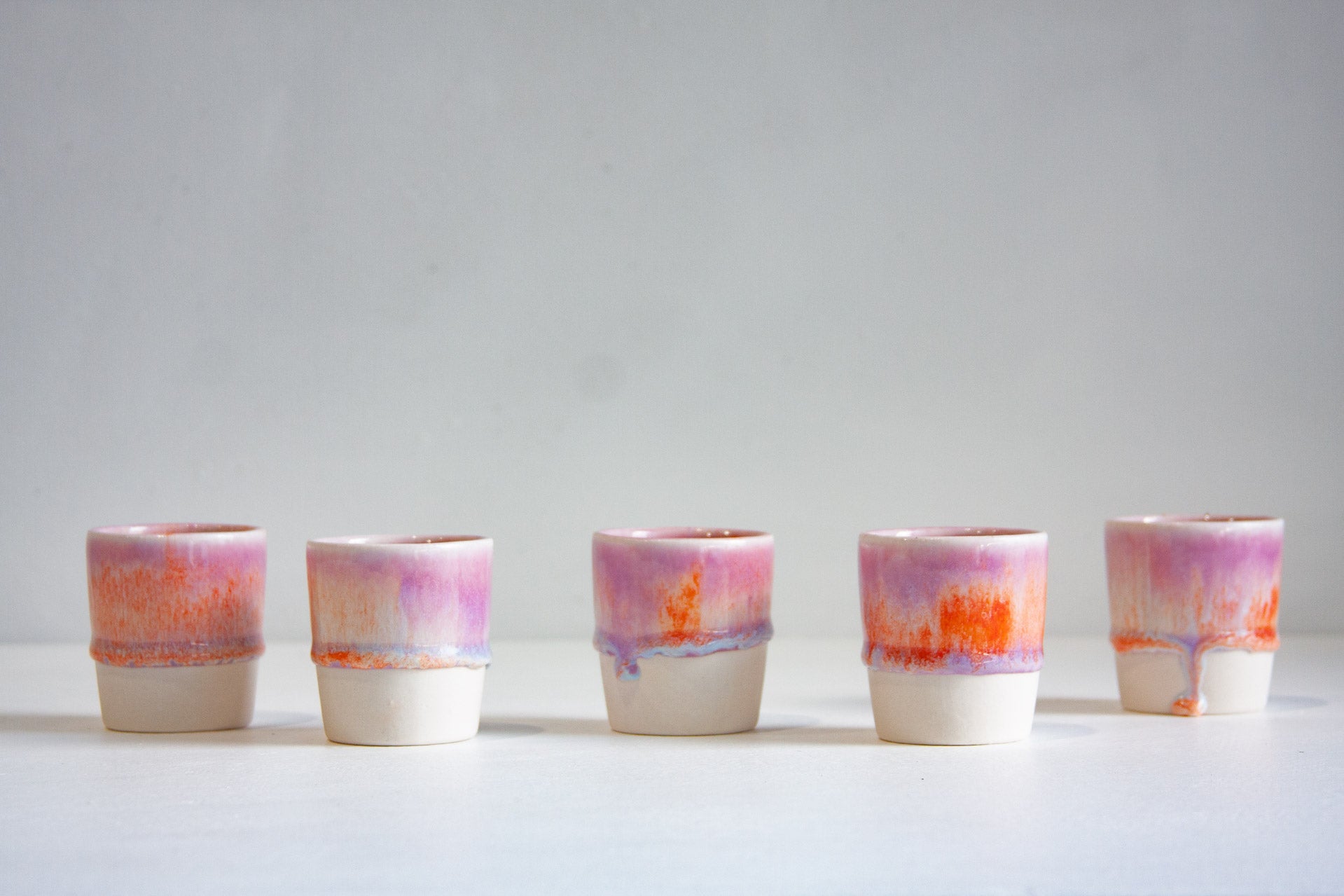 Five small ceramic tumblers with a two-tone design, featuring a white base and a top section glazed in pink, purple, and orange, arranged in a row against a light gray background.