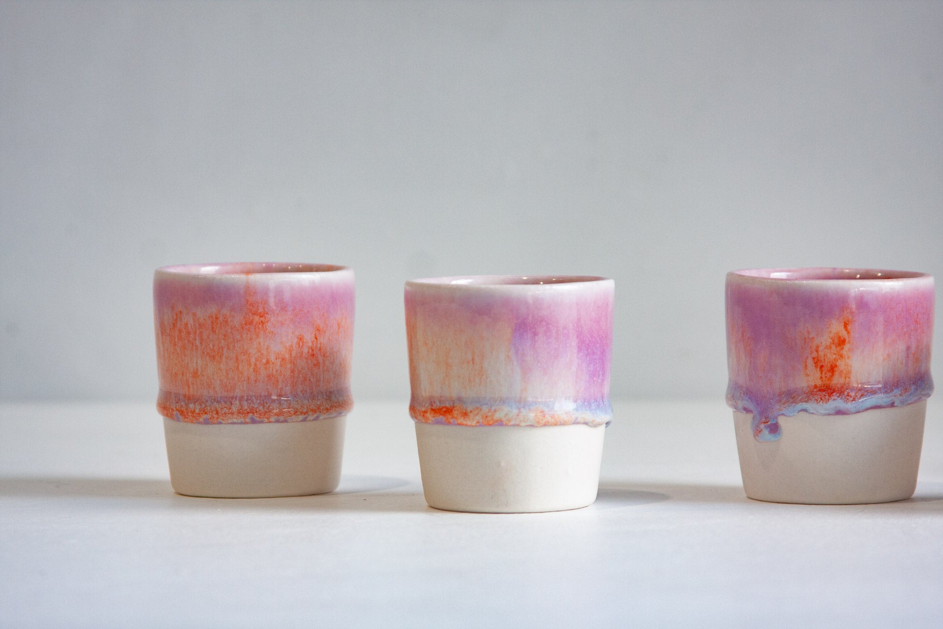 Three small ceramic cups with a pink, purple, and orange marbled glaze on the upper half and a matte off-white base, arranged on a light gray surface against a neutral gray background.