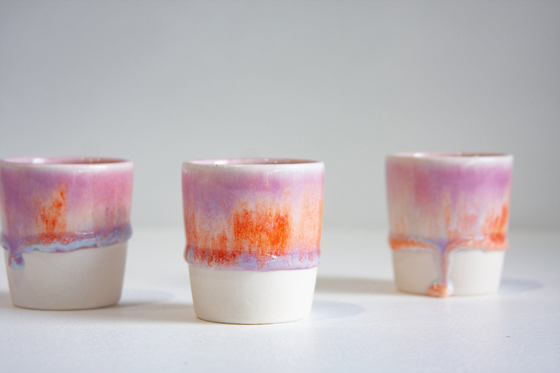 Three small ceramic cups with a white base and a glazed upper section featuring pink, purple, and orange abstract dripping patterns, arranged on a light gray surface against a light gray background.