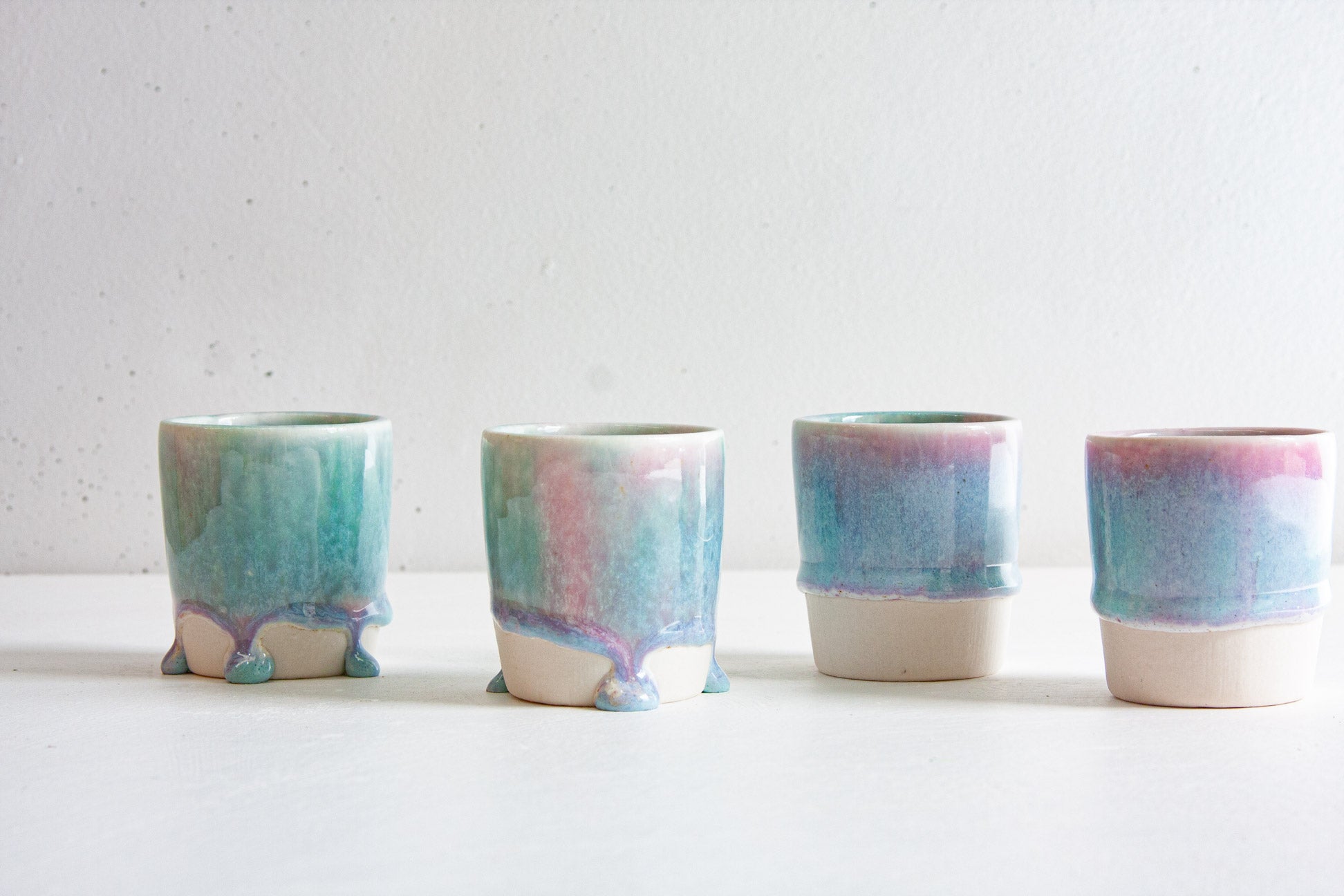 Four handmade ceramic espresso cups in a row, each in a light purple and blue glaze ending above the bare clay base, some running into drips, against a plain light background.