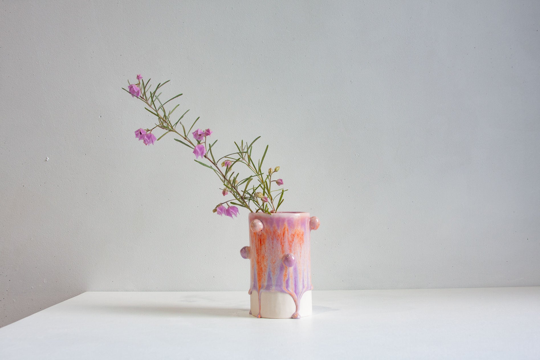 A small handmade ceramic vase with a pink, orange, and purple drip glaze, holding a delicate sprig of purple flowers, set on a white surface against a light gray background.