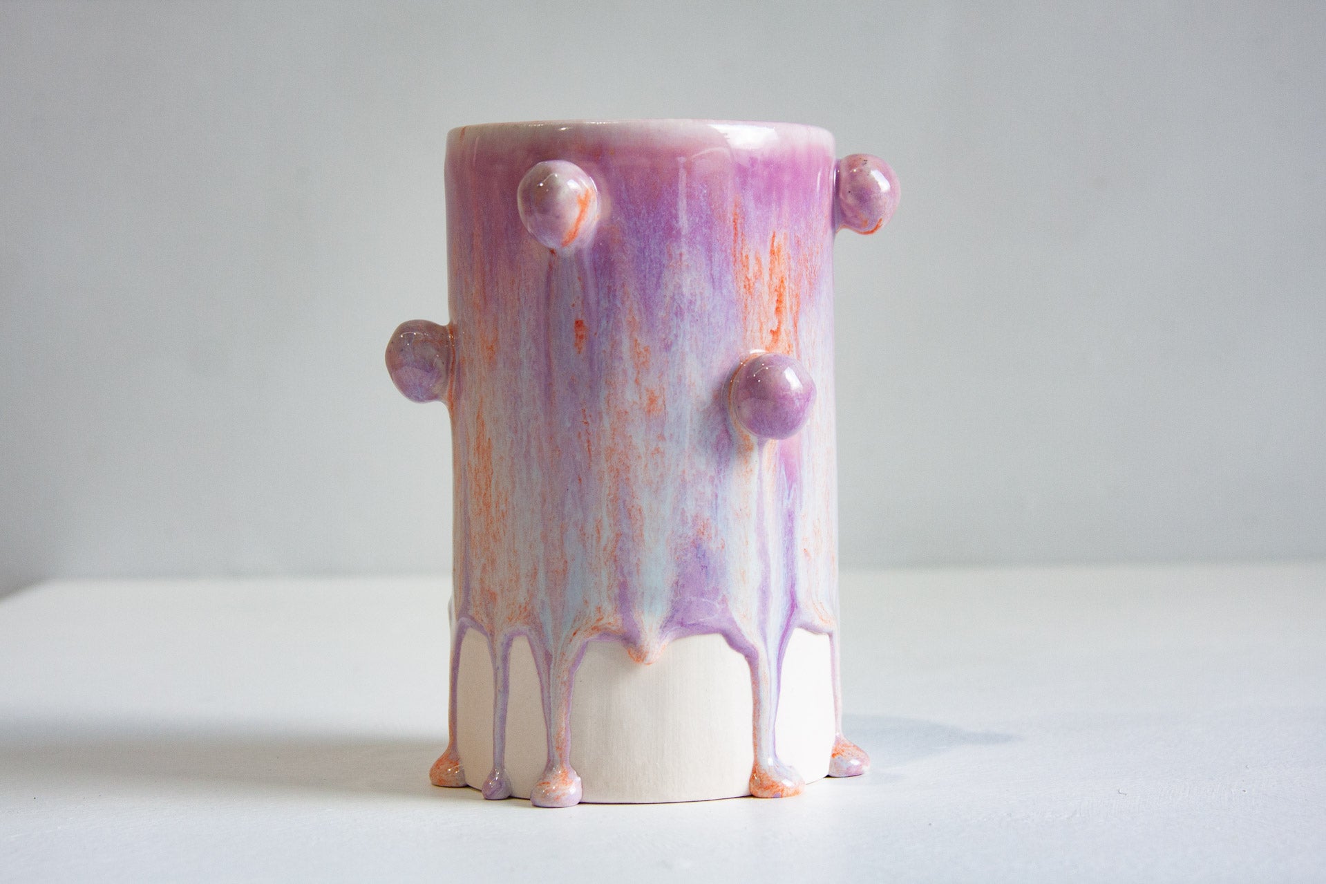 A handmade ceramic pot with a dripping paint design in shades of purple, pink, and orange, featuring small protruding bumps on the sides, set against a plain light grey background.