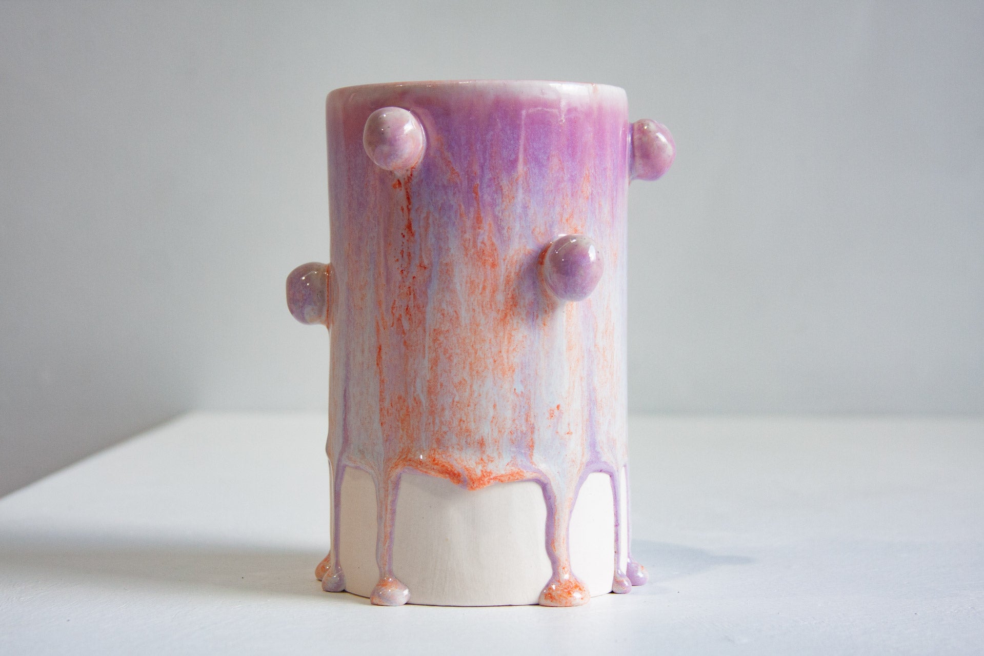 A handmade ceramic cylinder vase with a dripping paint design in shades of purple, pink, and orange, featuring small protruding spherical knobs, set against a plain light grey background.