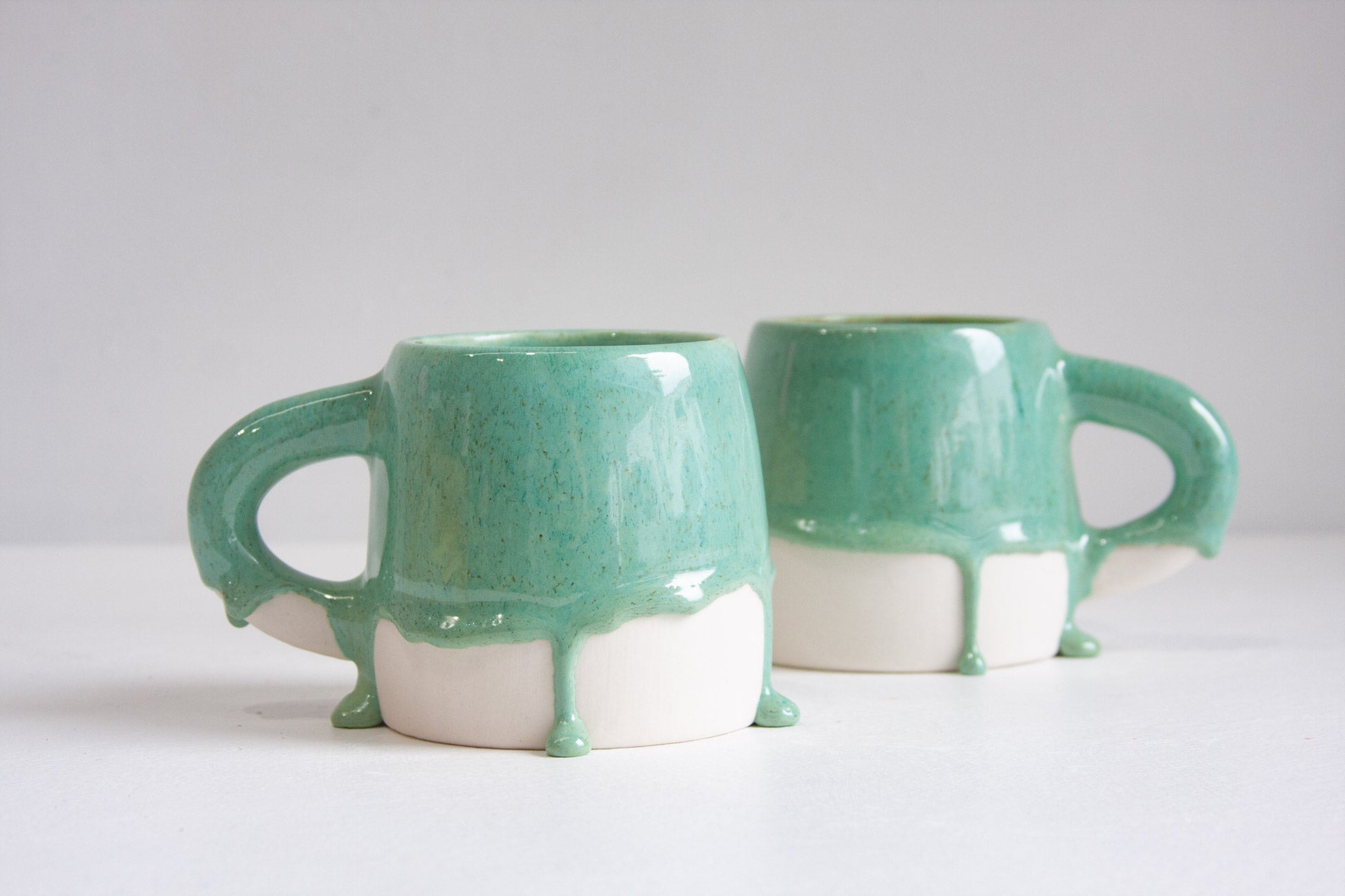 Two ceramic mugs with a glossy mint green and white dripping glaze design, displayed on a clean white surface against a light gray background.