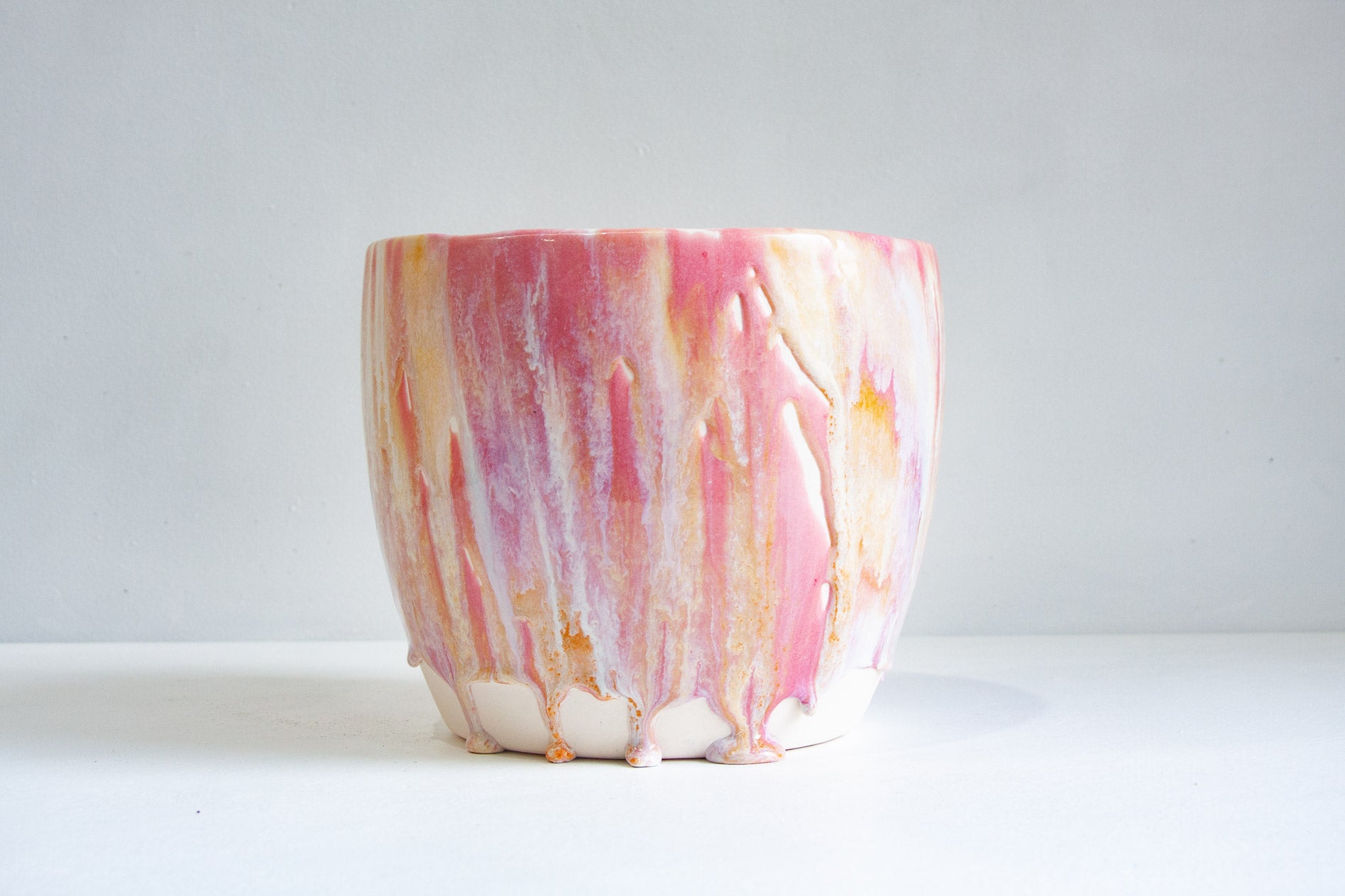 A handmade ceramic planter with a textured, dripping glaze finish in pink, white, and light orange, set against a plain light gray background.