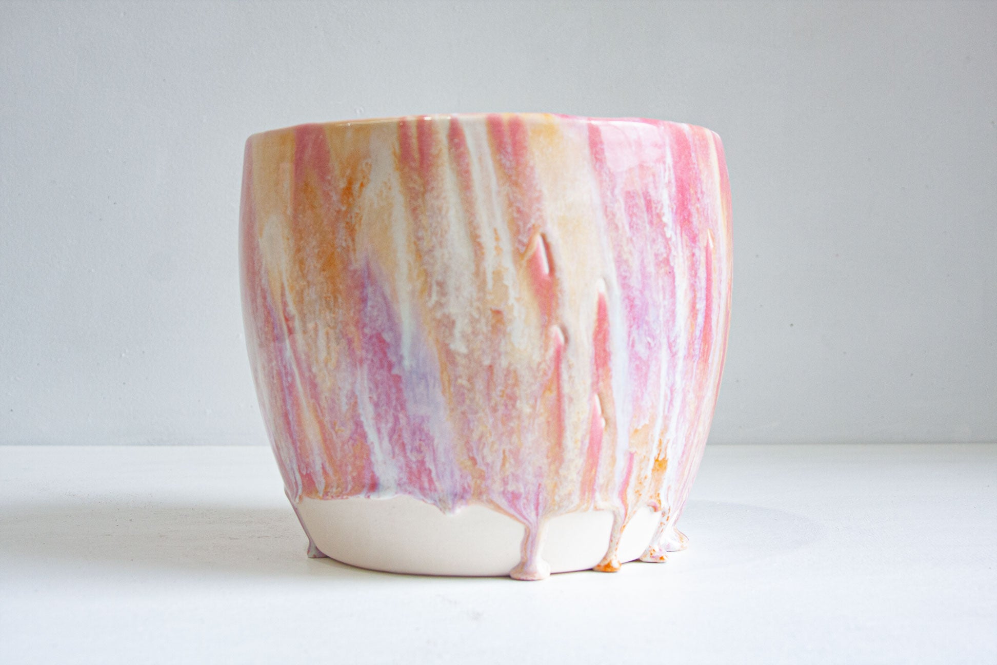 A ceramic planter with a marbled drip glaze design in pink, orange, yellow, and white, sitting on a white surface against a light gray background.