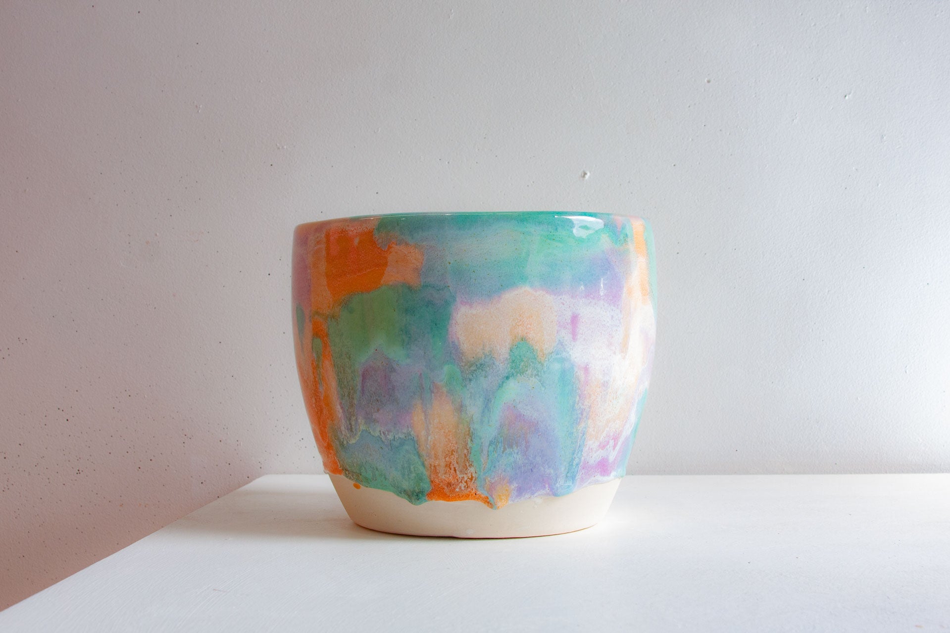 A handmade ceramic planter photographed front on, the pastel purple, peach and green glaze fading into the bare white clay at the foot.