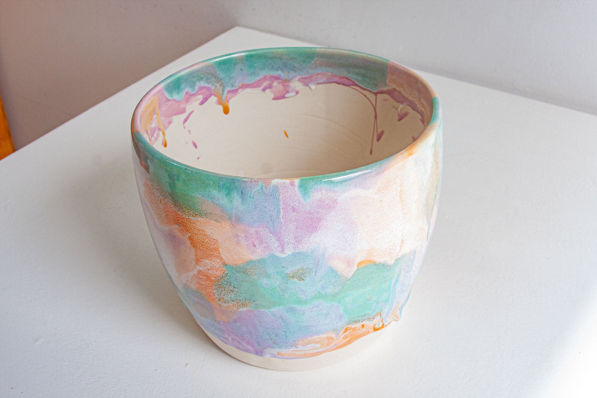 A handmade ceramic planter seen from above at an angle, showing the white glazed interior and pastel purple, peach and green washes running down the outside.