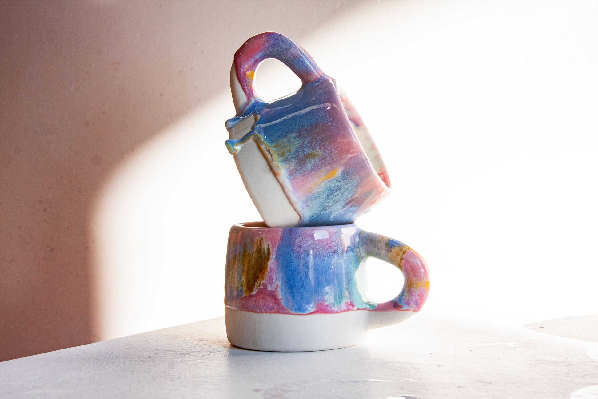 Two stacked ceramic mugs with colorful, painterly blue, pink, purple, and yellow glazes on a light gray surface against a warm beige wall with soft sunlight.