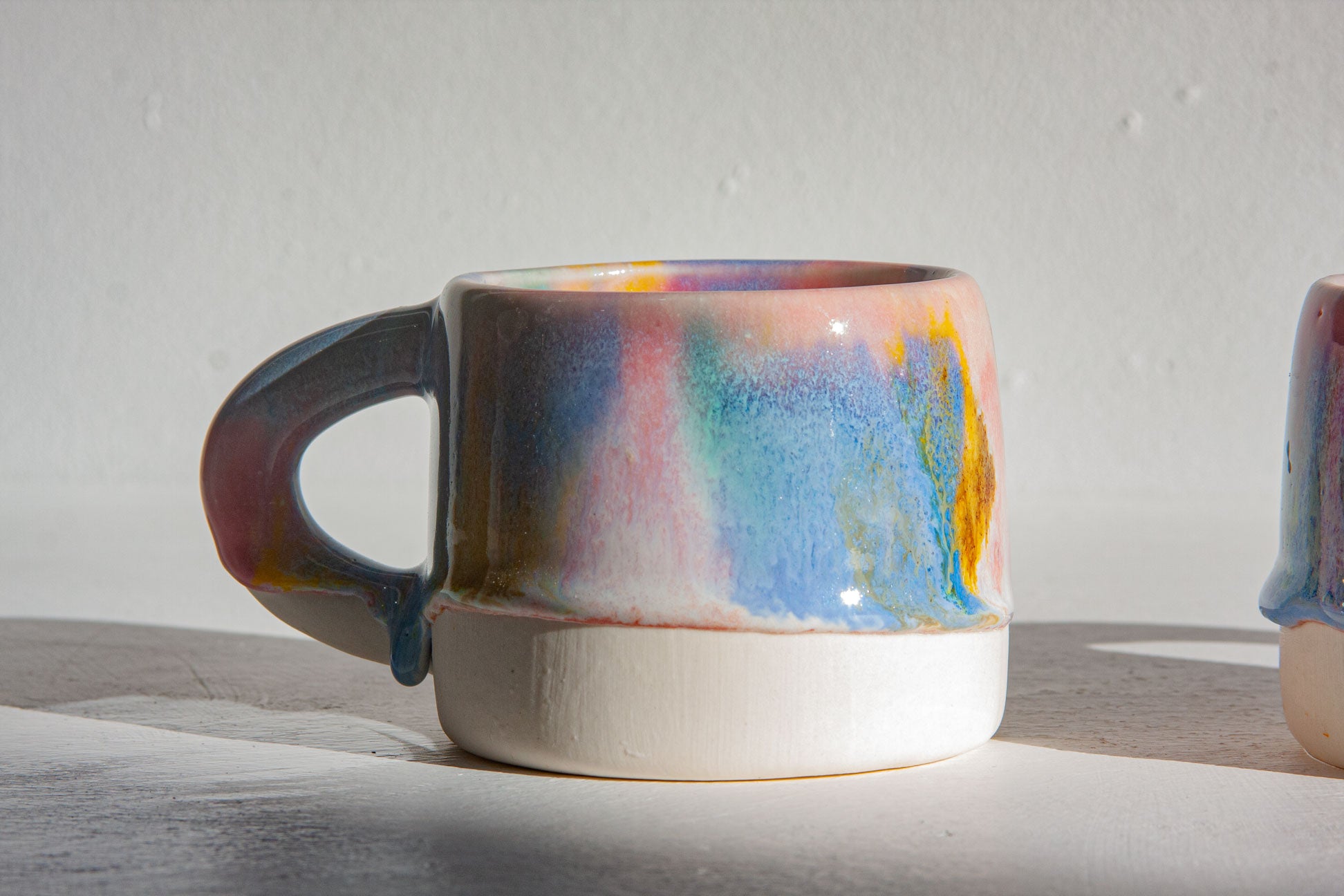 A handmade ceramic mug with a multicolored pastel glaze featuring blue, pink, yellow, and orange tones over a white base, set against a neutral light gray background.