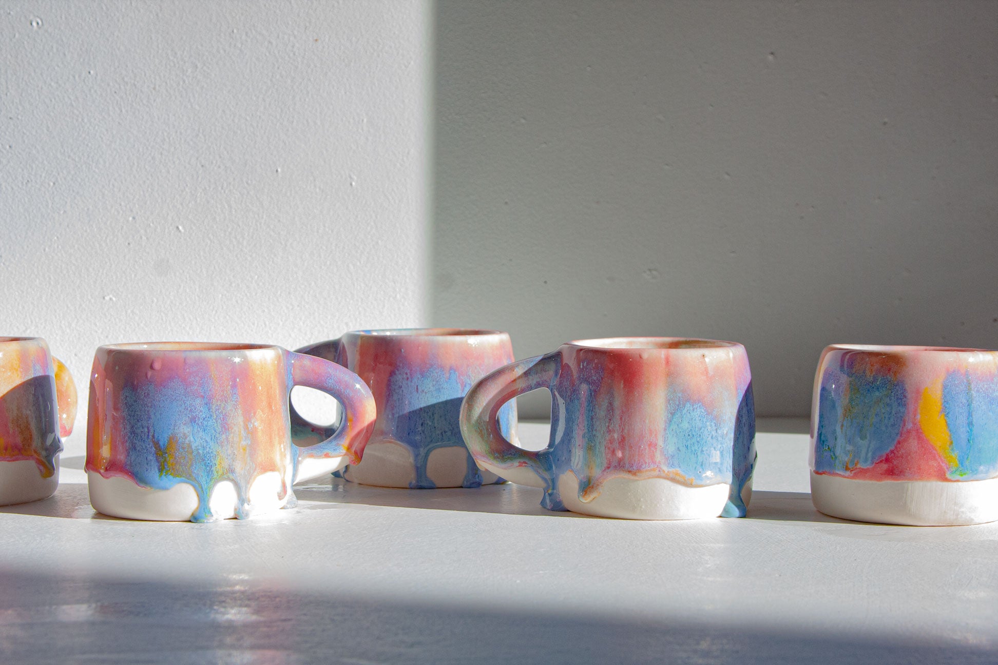 A row of small ceramic mugs with colorful drip glazes in pink, blue, purple, and yellow on a white surface against a light gray wall.